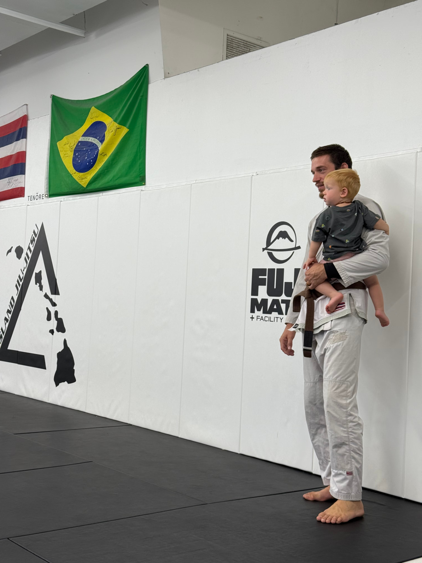 Coach holding his son on the mats beside the Island Jiu-Jitsu triangle logo